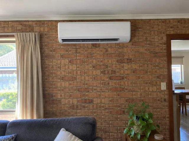 White split-system indoor unit on a brick wall in a living room with curtains and sofa in Andrews Farm